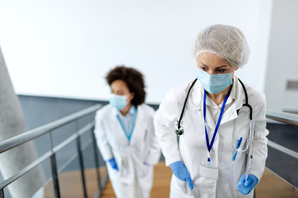 Worried female doctor wearing protective face mask and walking upstairs in a hallway at medical clinic. Her colleague is in the background.